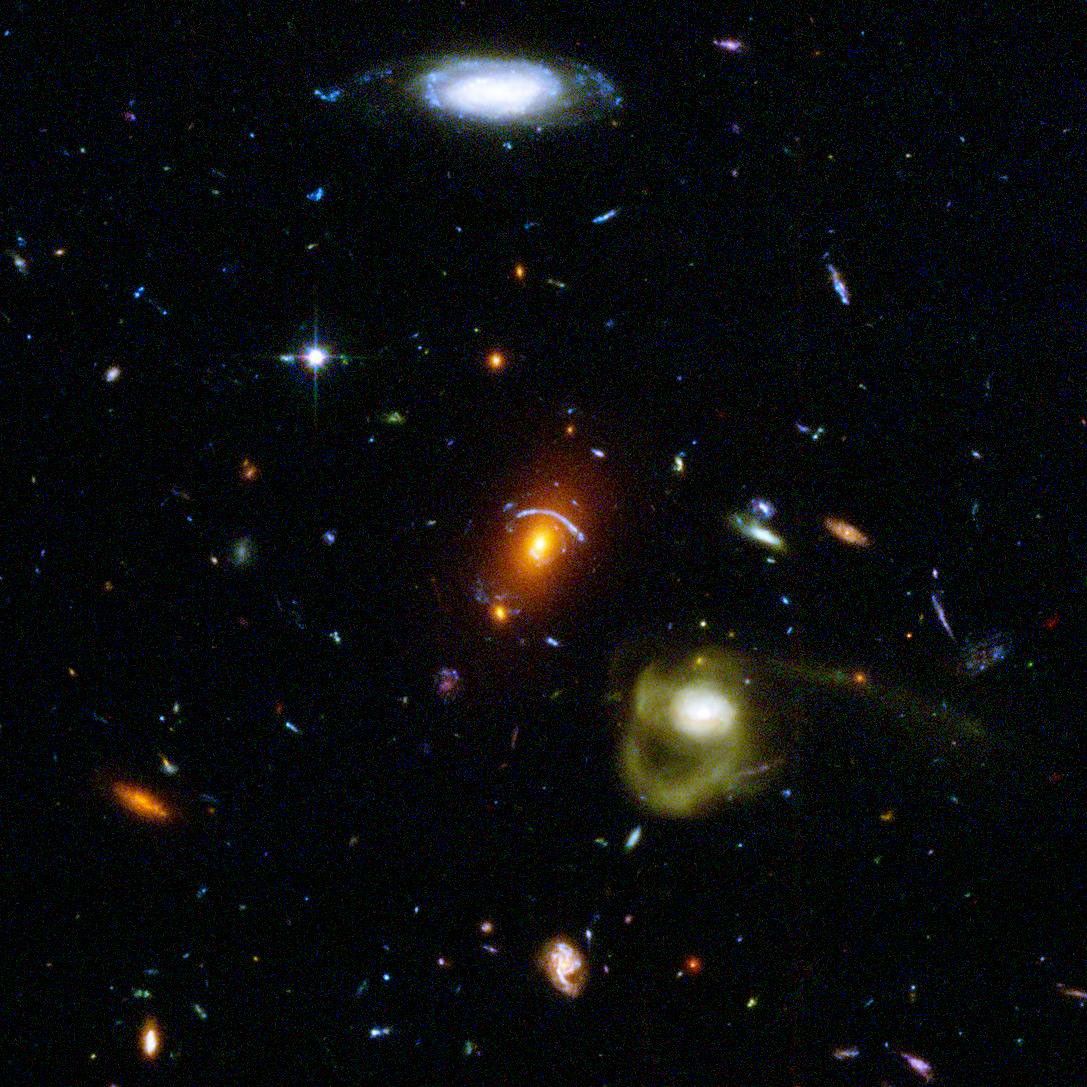 Black background dotted with galaxies. A reddish galaxy at image center with a bluish-white arc above it. Below it to the right it a yellowish swirling galaxy, and at the top center is a blue-white spiral galaxy. Other spiral and elliptical galaxies appear smaller throughout the image, as they are farther away.